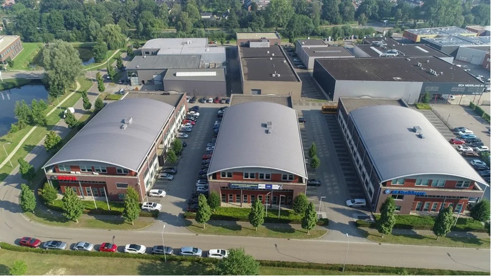 Luchtopname van drie kantoorpanden aan De Wel in een bedrijventerrein met omliggende parkeerplaatsen en groen.